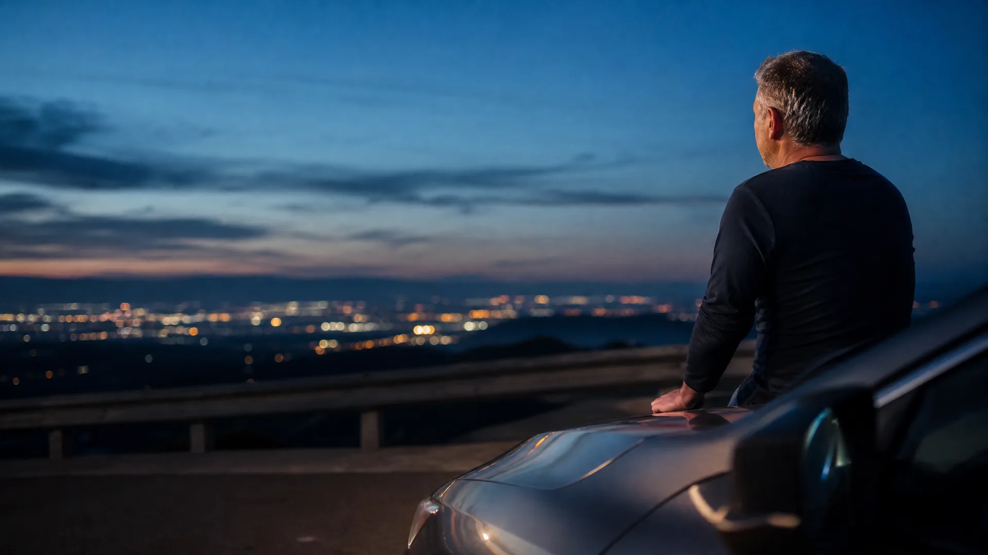 A man in a dark long-sleeved shirt leans against the hood of a car at dusk, looking out over a distant city's lights beneath a deep blue sky.