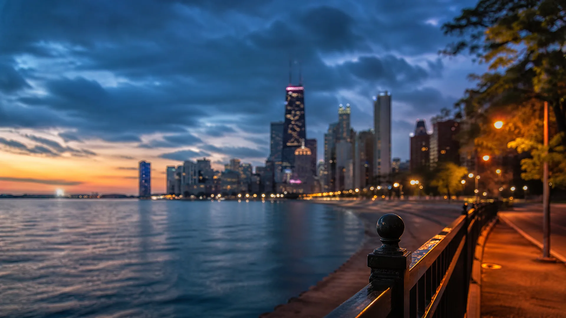 A city skyline of tall lit buildings across a large body of water at dusk, seen past a dark metal railing with a rounded post on a curving waterfront path.